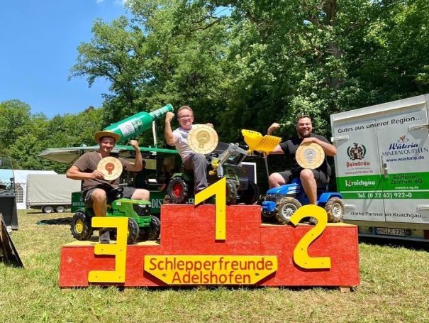 Drei Männer auf Podest mit Pokalen, umgeben von Traktoren in einem grünen Umfeld.